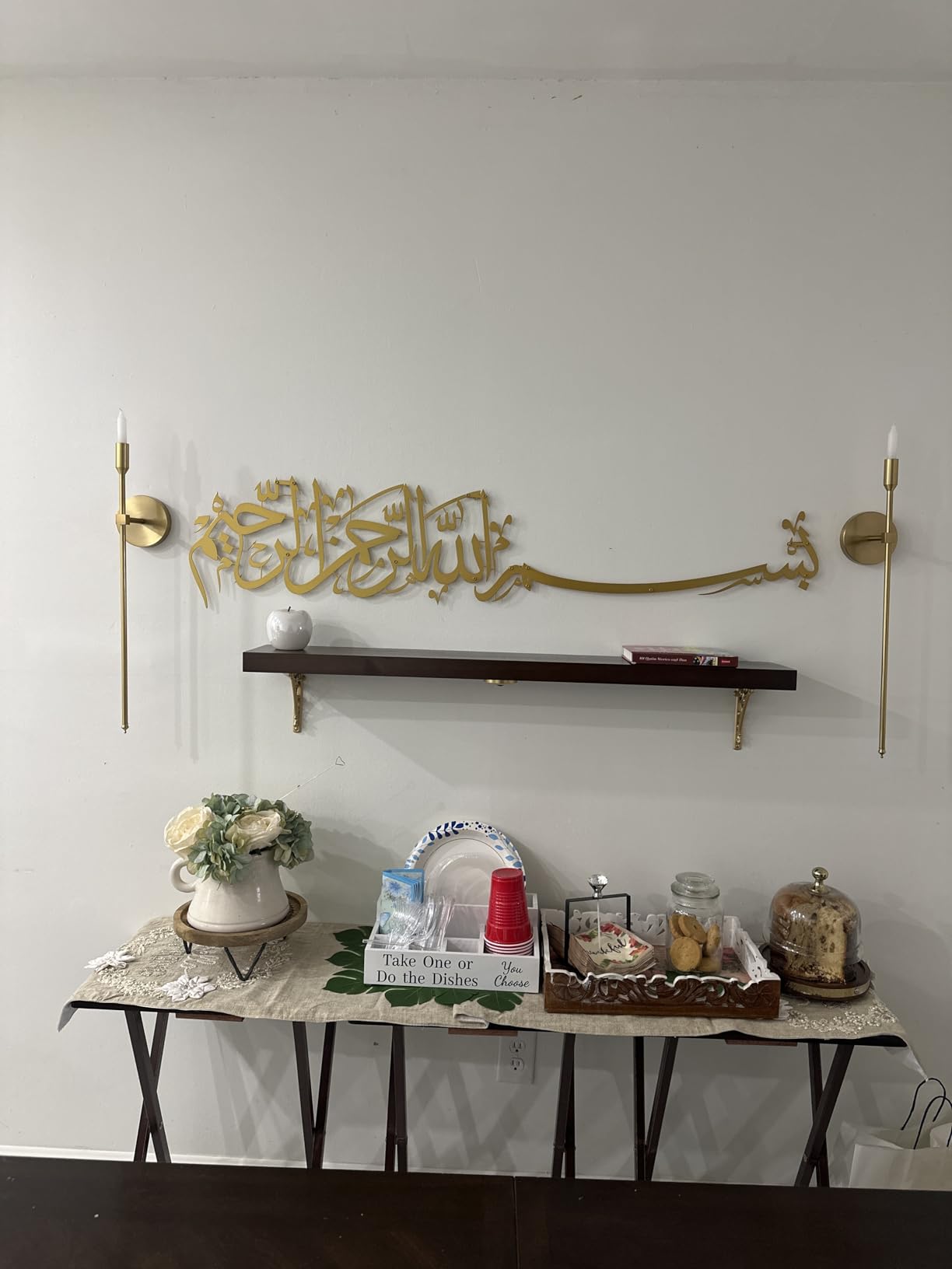 Decorative wall shelf with gold Arabic calligraphy, candles, and small items on a white wall.