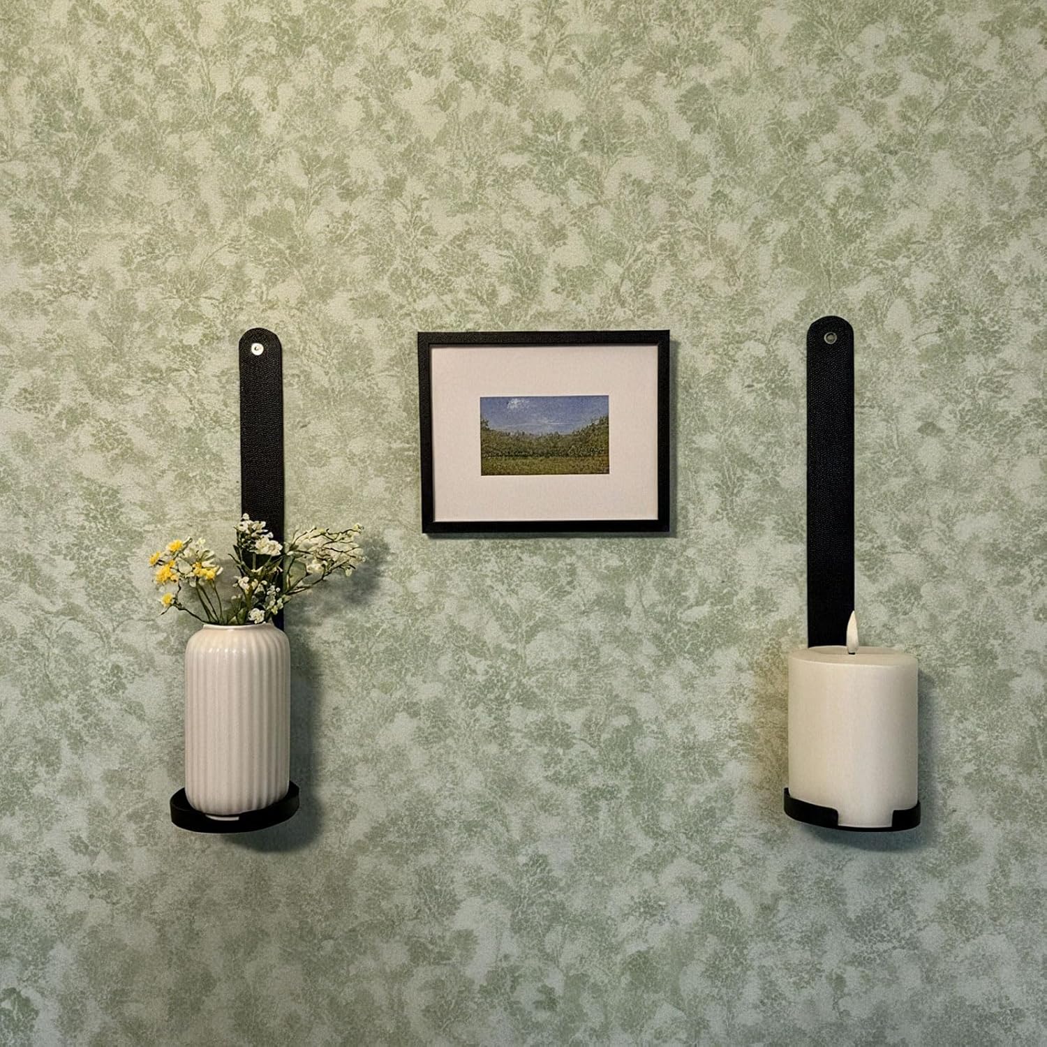 Decorative wall with a framed picture, a vase with flowers, and two candle holders against a patterned wall.