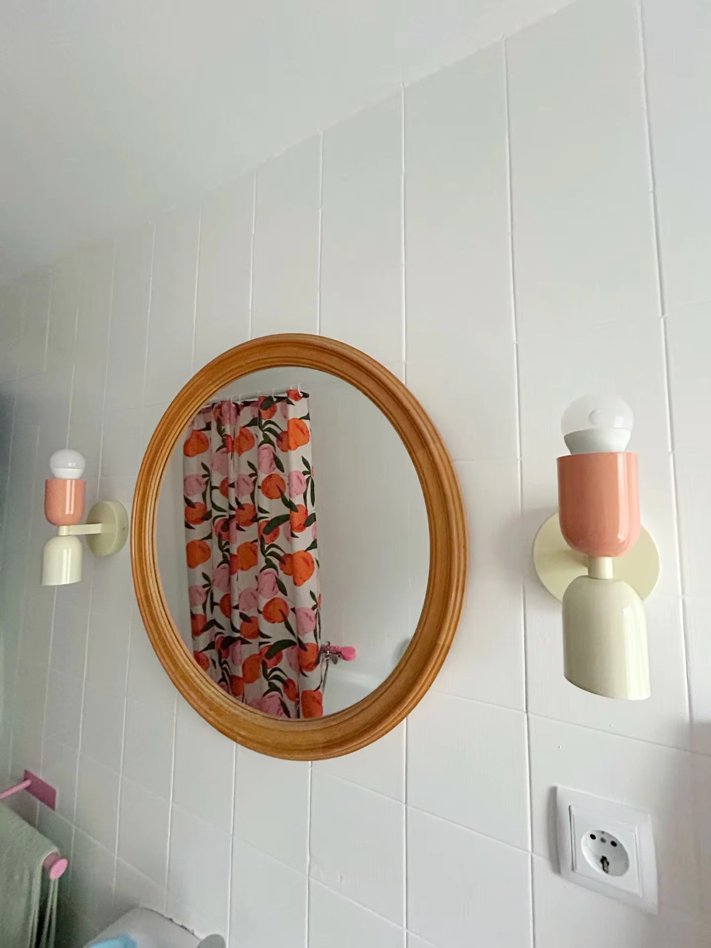 Round wooden mirror on a white tiled wall with two pink sconces.