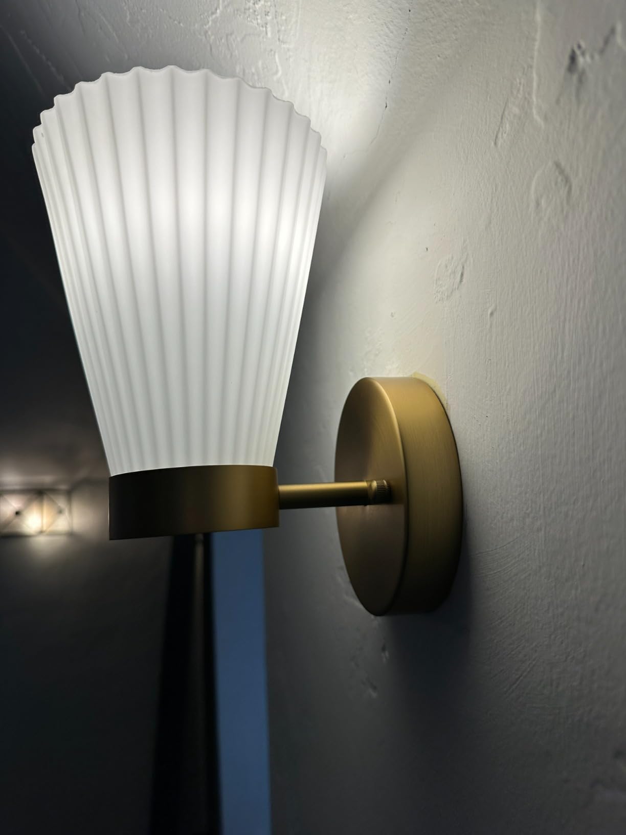 Wall-mounted light fixture with white ribbed shade and gold fixture on a gray wall.